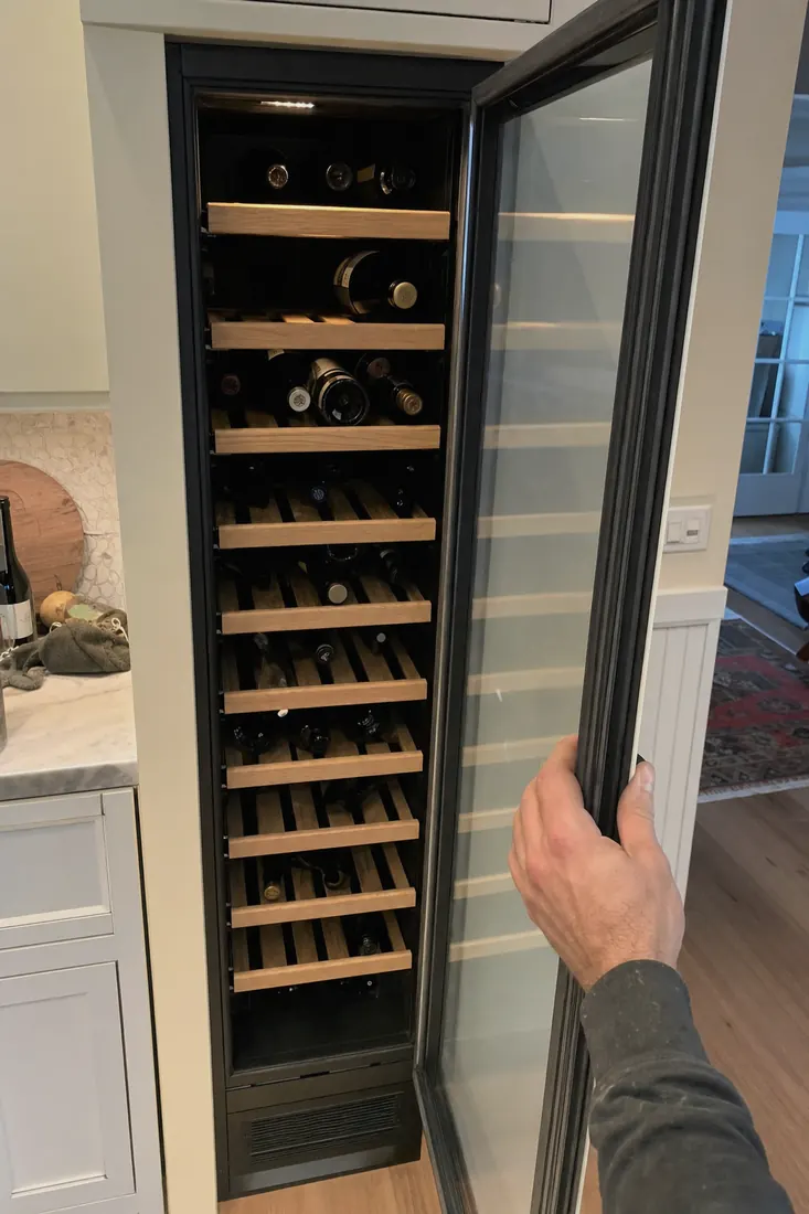 An open built-in wine column, bottles on wooden shelves, a hand at the door seal - Accessibility