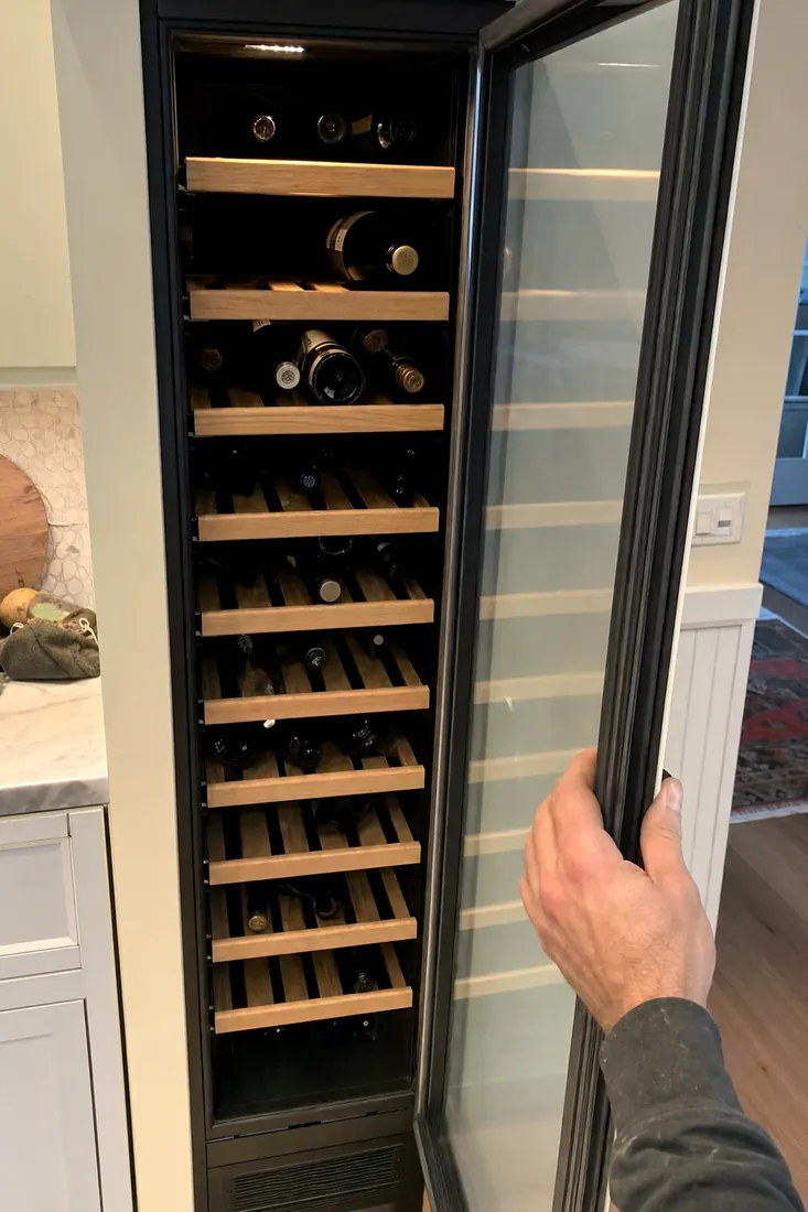 An open built-in wine column, bottles on wooden shelves, a hand at the door seal, the surrounding cabinetry in frame - Wine Cooler Not Holding Temperature?