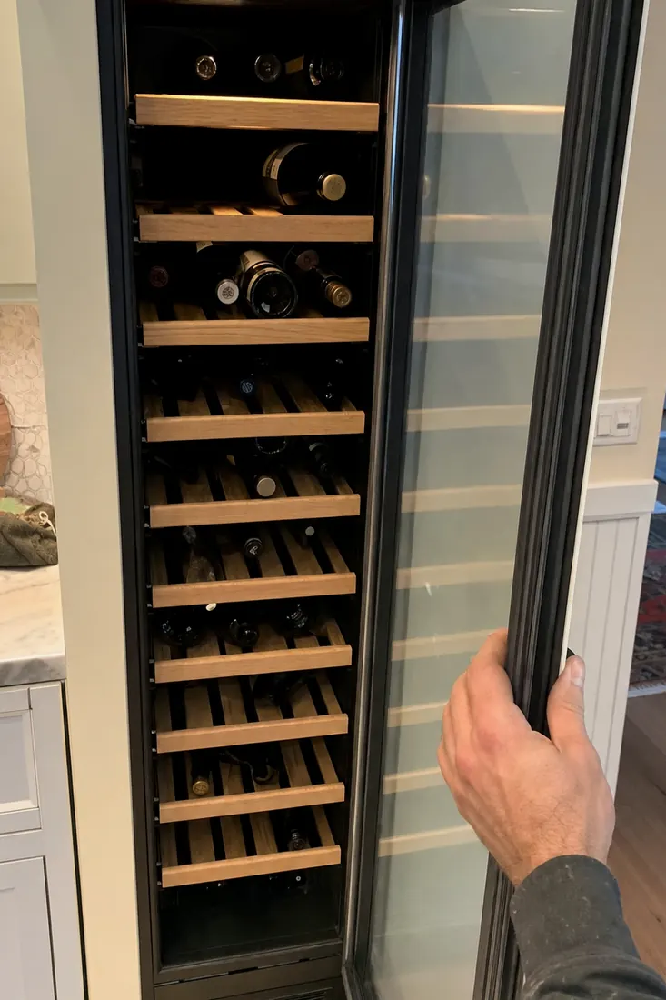 An open built-in wine column, bottles on wooden shelves, a hand at the door seal, the wooden shelves visible in the picture - Sub-Zero and luxury appliance repair in Atlantic Beach