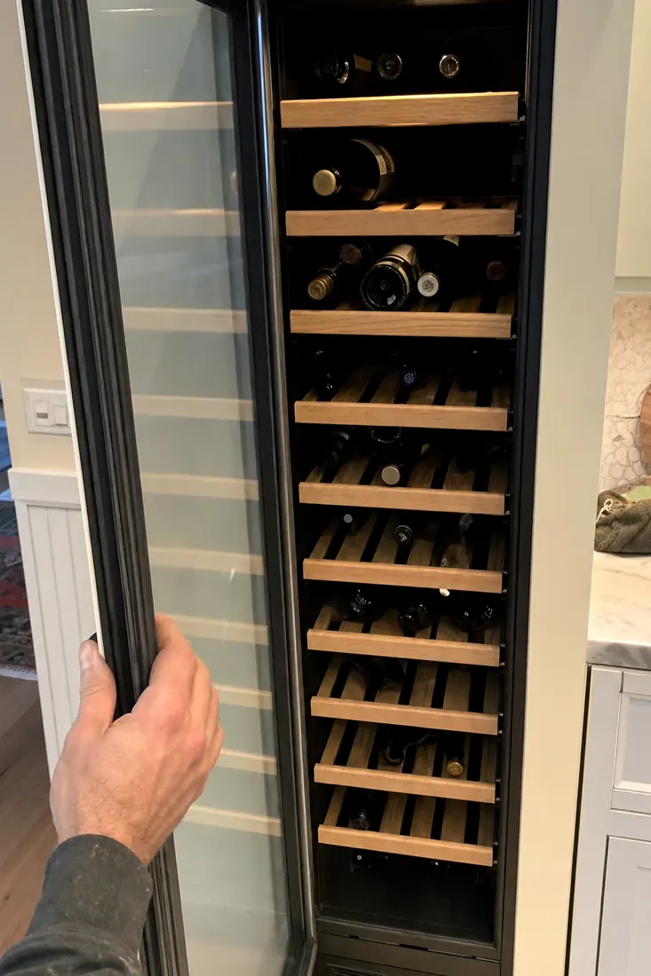 An open built-in wine column, bottles on wooden shelves, a hand at the door seal, the surrounding cabinetry visible in the picture - Sub-Zero and luxury appliance repair in Durbin Crossing