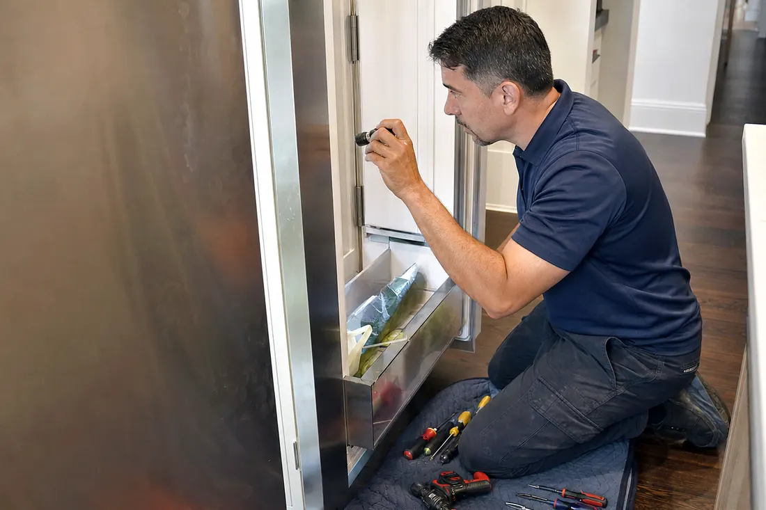 Technician checking inside an open built-in refrigerator with a flashlight, the kitchen floor visible in the picture - Maintenance