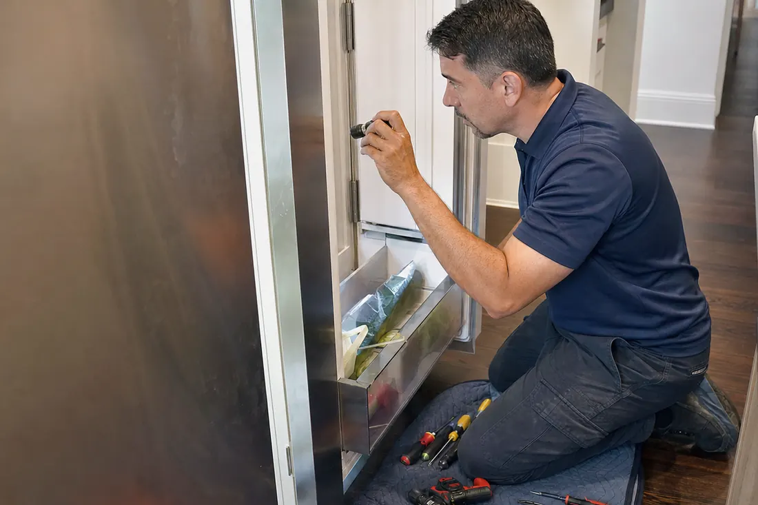 Technician checking inside an open built-in refrigerator with a flashlight, the open door in frame - How to Clean a Sub-Zero Condenser