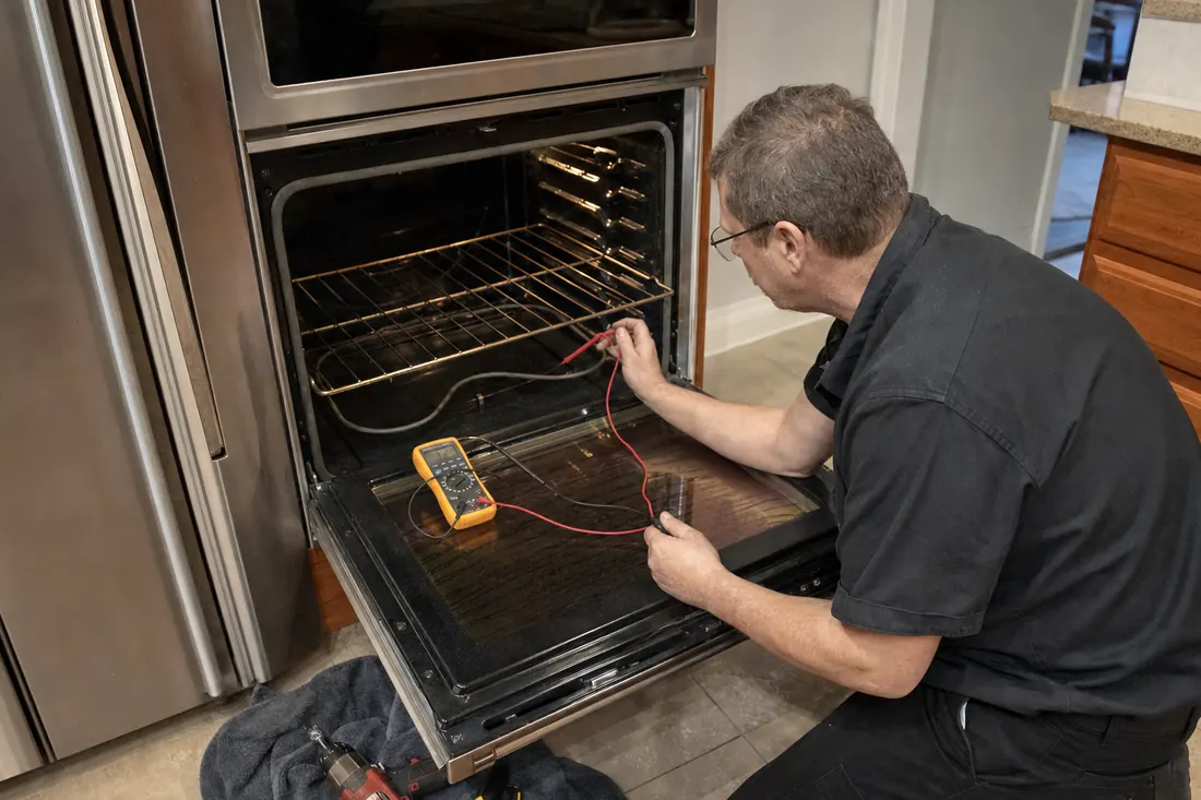 Technician testing the bake element of an open double wall oven with a multimeter, tools on a towel on the tile floor in frame - Straight answers