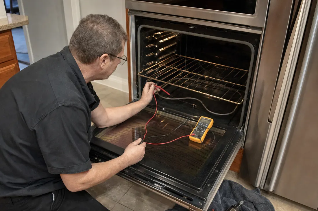 Technician testing the bake element of an open double wall oven with a multimeter, tools on a towel on the tile floor visible in the picture - What to check before you call anyone